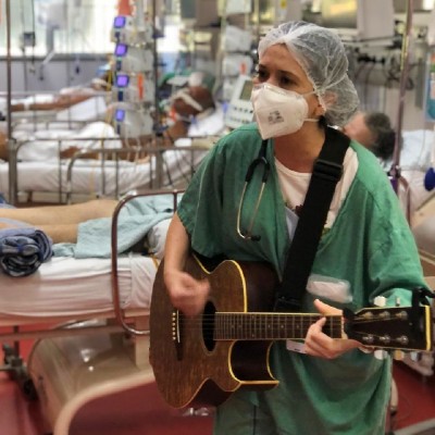 Foto de Bruna Falluh Alarcão. Ela está com um uniforme verde, touca e máscara, dentro de uma UTI, tocando violão para os pacientes internados